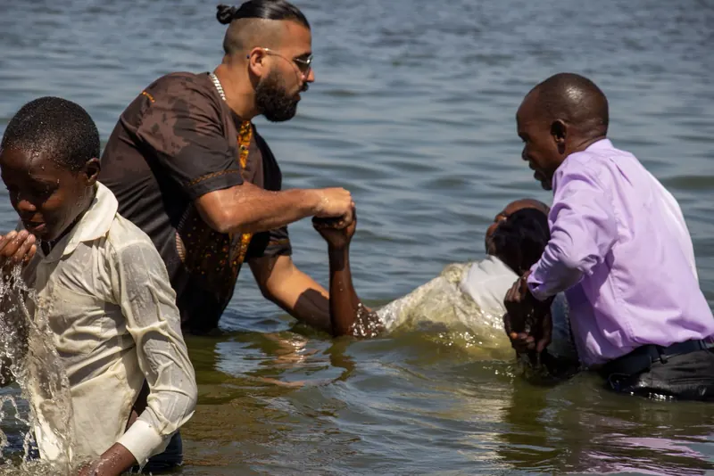 A baptism in open water: two men lower a person into the water, with a boy beside them