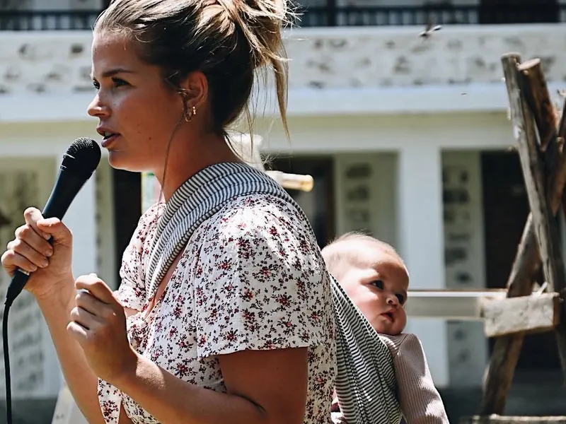A woman speaks into a microphone with a baby carried on her back