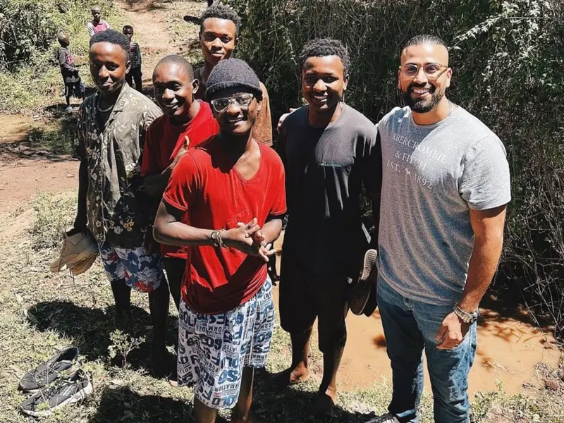 Six men stand together on a dirt path, with children in the distance behind them